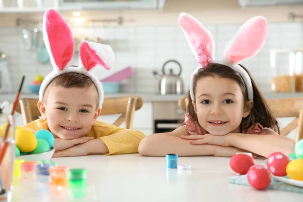 Милые дети с повязками на ушах и раскрашенными пасхальными яйцами, сидящие за столом на кухне — стоковое фото