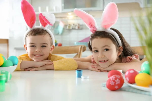 Niedliche Kinder mit Hasenohren-Stirnbändern und bemalten Ostereiern sitzen am Tisch in der Küche — Stockfoto