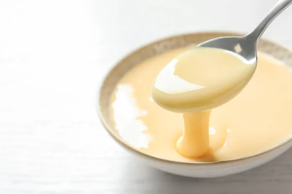 Spoon of pouring condensed milk over bowl on table, closeup with space for text. Dairy products