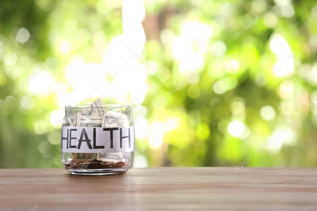 Glass jar with money and word HEALTH on table against blurred background, space for text