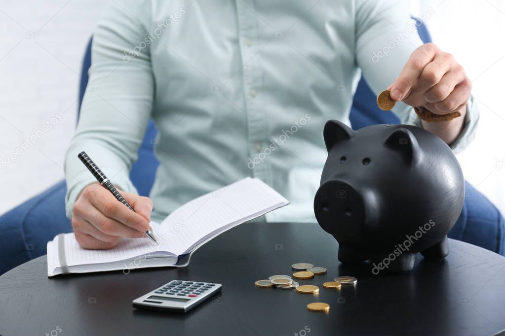 Businessman with notebook, piggy bank and money at table against light background, closeup