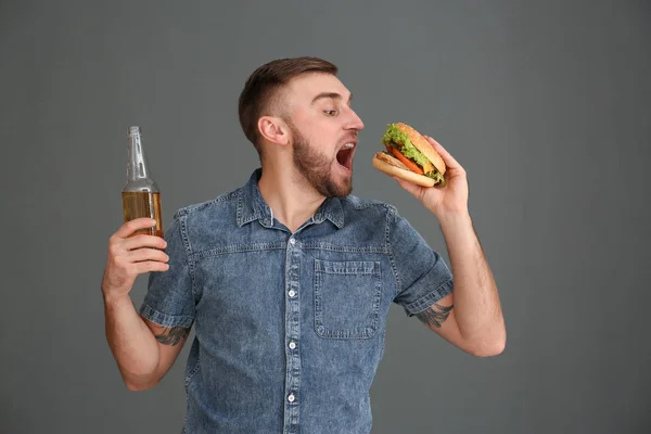 Ung man med öl äta välsmakande hamburgare på grå bakgrund — Stockfoto