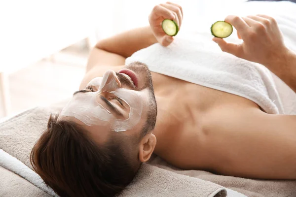 Giovane uomo con maschera facciale che tiene fette di cetriolo nel salone spa — Foto Stock