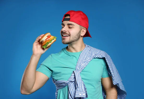 Handsome man eating tasty burger on color background — Stock Photo, Image