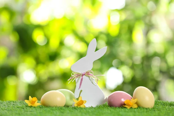 Cute wooden Easter bunny and dyed eggs on green grass against blurred background — Stock Photo, Image