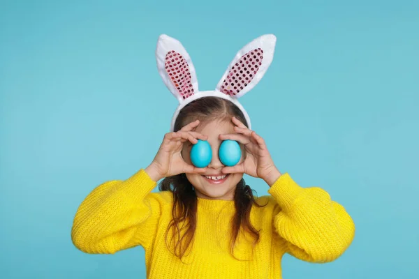 Niña en orejas de conejo diadema sosteniendo huevos de Pascua cerca de los ojos en el fondo de color — Foto de Stock