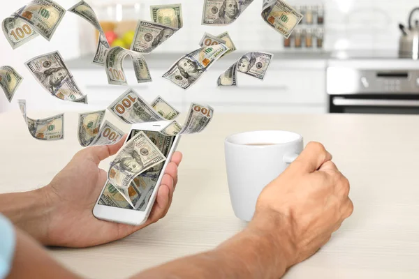 Человек с мобильным телефоном и летающими долларовыми банкнотами за столом на кухне, крупным планом. Люди зарабатывают деньги в интернете — стоковое фото