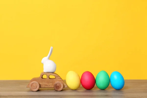 Toy car with ceramic bunny and Easter eggs on wooden table against color background — 图库照片