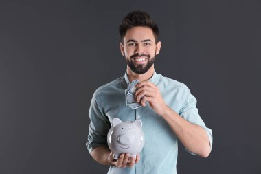 Mutlu genç adam kumbara gri arka plan üzerinde içine para koyarak