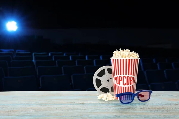 Copa de palomitas de maíz, carrete y vasos sobre mesa de madera en sala de cine vacía. Espacio para texto — Foto de Stock