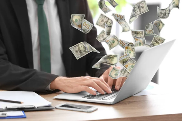 Человек с современным ноутбуком и летающими долларовыми банкнотами за столом в офисе, крупным планом. Люди зарабатывают деньги онлайн за столом — стоковое фото