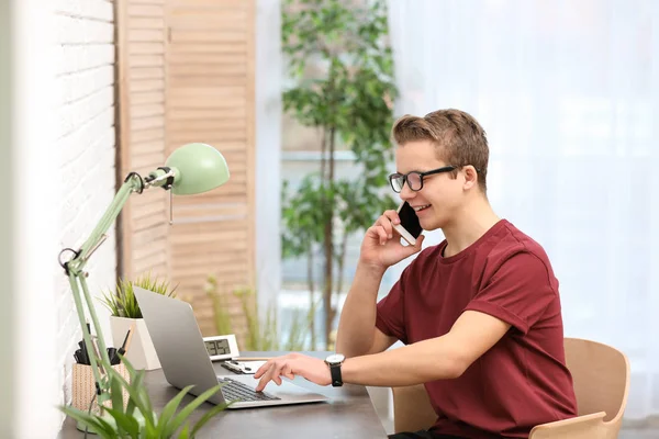 Knappe tiener praten over telefoon en met behulp van laptop aan tafel in de kamer — Stockfoto