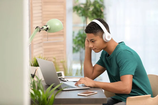 Adolescente afroamericano con auriculares usando portátil en la mesa en la habitación —  Fotos de Stock