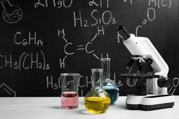 Εργαστηριακά γυαλικά και μικροσκόπιο στο τραπέζι ενάντια σε μαυροπίνακα με χημικούς τύπους — Φωτογραφία Αρχείου
