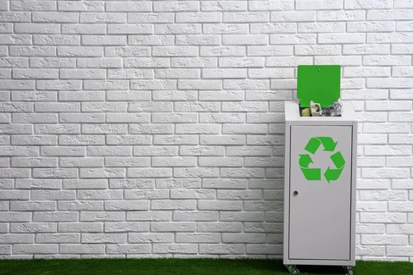 Overfilled trash bin with recycling symbol near brick wall indoors. Space for text