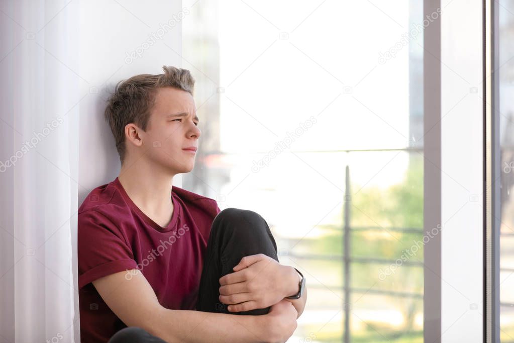 Upset teenage boy sitting alone near window indoors