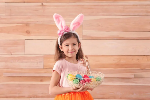 Little girl in bunny ears headband holding basket with Easter eggs against wooden background, space for text — Stock Photo, Image