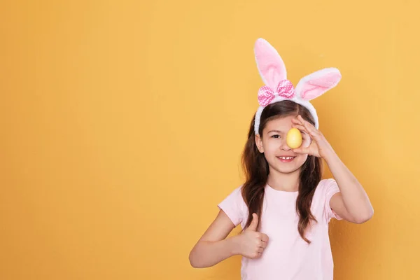 Niña en orejas de conejo diadema celebración de huevo de Pascua cerca de ojo sobre fondo de color, espacio para el texto — Foto de Stock