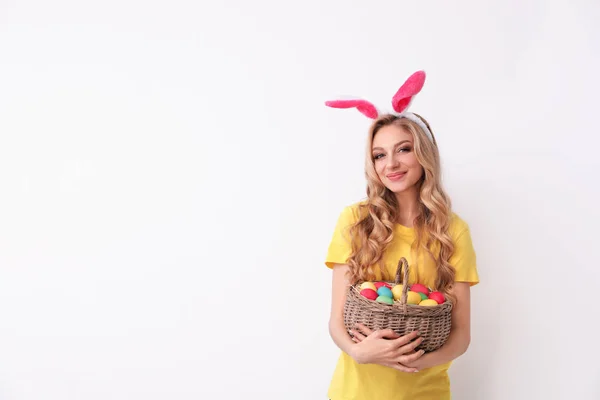 Beautiful young woman in bunny ears headband holding basket with Easter eggs on light background, space for text — Stock Photo, Image