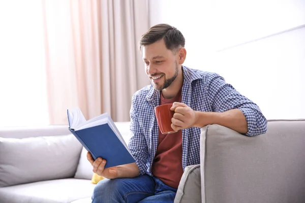 Schöner Mann mit einer Tasse Kaffee beim Lesen eines Buches auf dem heimischen Sofa — Stockfoto
