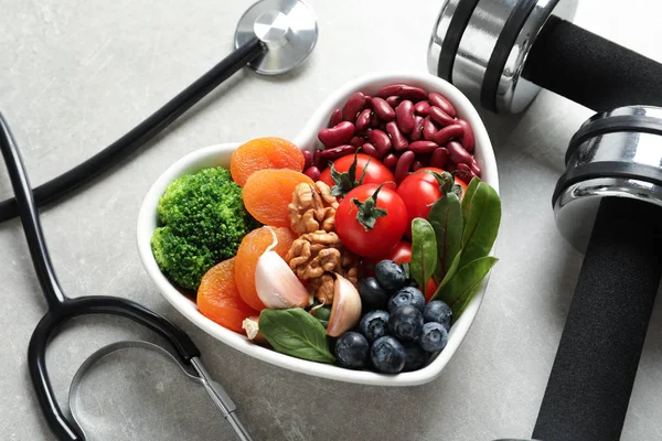 Cuenco de productos para una dieta saludable para el corazón, pesas y estetoscopio sobre una mesa gris — Foto de Stock