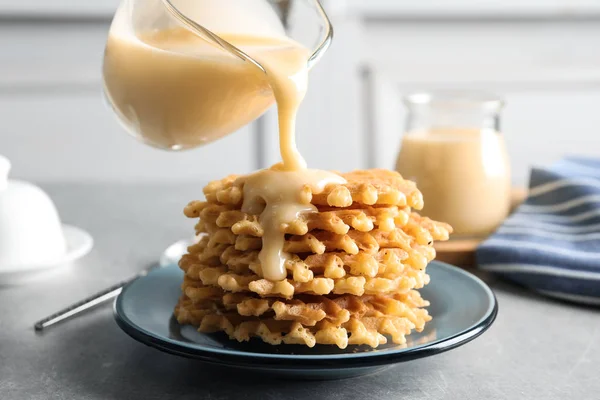 Pouring condensed milk from jug onto waffles on grey table. Dairy product