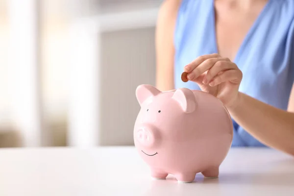 Vrouw zetten munt in Piggy Bank aan tafel binnenshuis, close-up. Ruimte voor tekst — Stockfoto