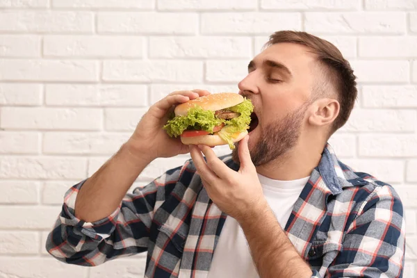 Jovem comendo hambúrguer saboroso contra parede de tijolo — Fotografia de Stock