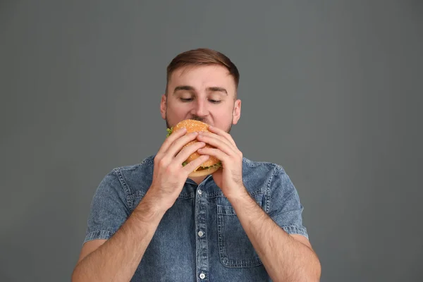 Young man eating tasty burger on grey background — Stock Photo, Image