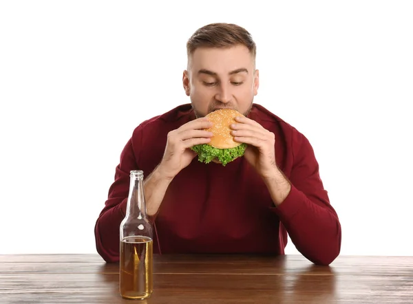 Jovem com cerveja comendo hambúrguer saboroso na mesa no fundo branco — Fotografia de Stock