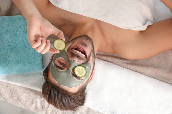 Young man with clay mask on his face holding cucumber slices in spa salon, above view — Stock Photo, Image