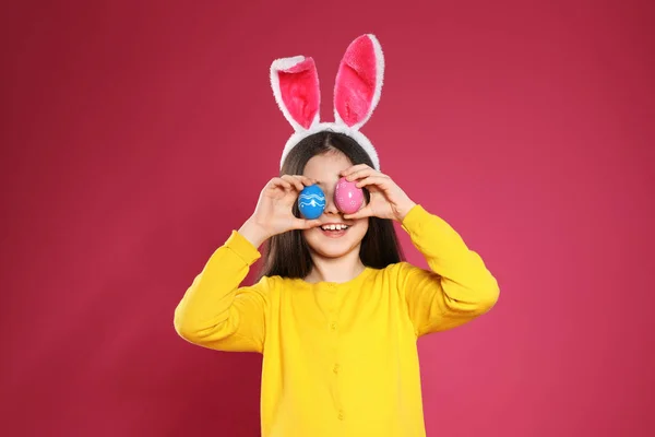 Niña en orejas de conejo diadema sosteniendo huevos de Pascua cerca de los ojos en el fondo de color — Foto de Stock