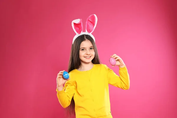 Little girl in bunny ears headband holding Easter eggs on color background — Stock Photo, Image