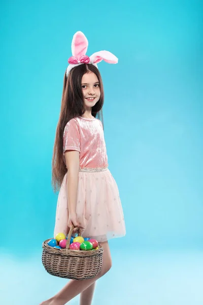 Little girl in bunny ears headband holding basket with Easter eggs on color background — Stock Photo, Image