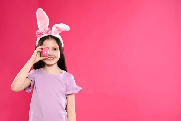 Little girl in bunny ears headband holding Easter egg near eye on color background, space for text — Stock Photo, Image