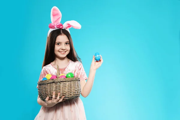 Little girl in bunny ears headband holding basket with Easter eggs on color background, space for text — Stock Photo, Image