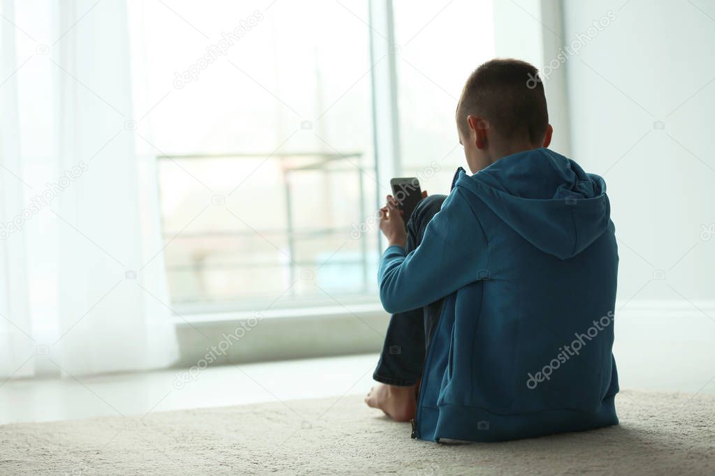 Upset boy with smartphone sitting near window indoors. Space for text