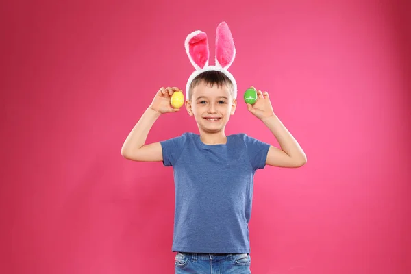 Kleiner Junge in Hasenohren Stirnband hält Ostereier auf farbigem Hintergrund — Stockfoto