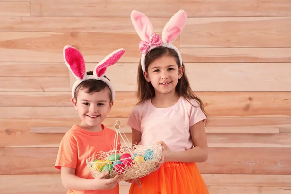 Lindos niños en orejas de conejo diademas celebración cesta con huevos de Pascua sobre fondo de madera — Foto de Stock