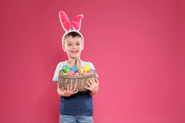 Menino em orelhas de coelho headband segurando cesta com ovos de Páscoa no fundo de cor, espaço para texto — Fotografia de Stock