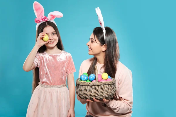 Mother and daughter in bunny ears headbands with basket of Easter eggs on color background — Stock Photo, Image