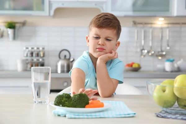 Adorable little boy refusing to eat vegetables at table in kitchen — Stock Photo, Image