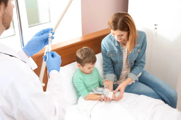 Médico ajustando gotejamento intravenoso para criança pequena no hospital durante a visita dos pais — Fotografia de Stock