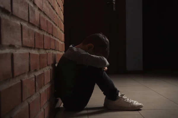 Triste niño sentado en el suelo cerca de la pared de ladrillo en el interior —  Fotos de Stock