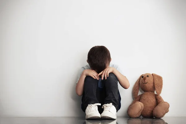 Petit garçon triste avec jouet assis près du mur blanc, espace pour le texte — Photo