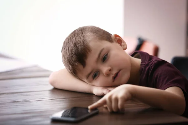 Triste petit garçon avec téléphone portable assis à la table à l'intérieur — Photo