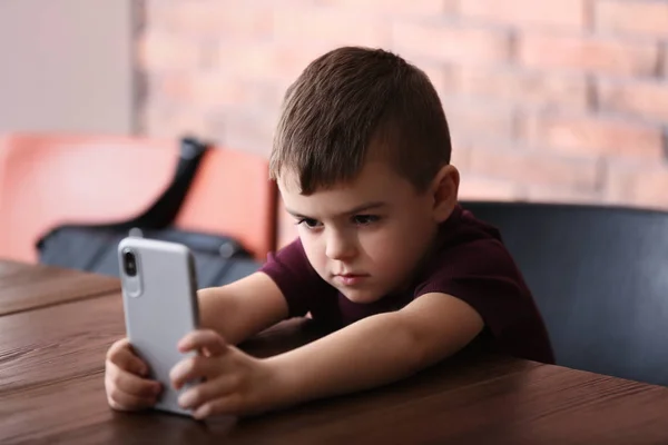 Triste petit garçon avec téléphone portable assis à la table à l'intérieur — Photo