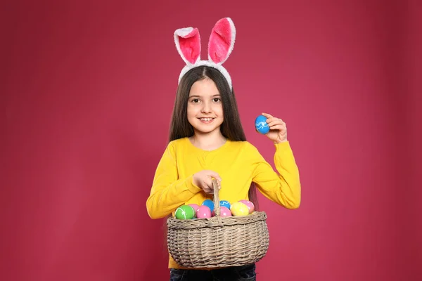 Little girl in bunny ears headband holding basket with Easter eggs on color background — Stock Photo, Image