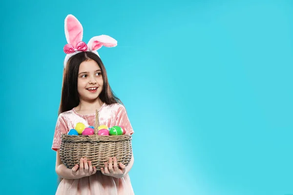 Little girl in bunny ears headband holding basket with Easter eggs on color background, space for text — Stock Photo, Image