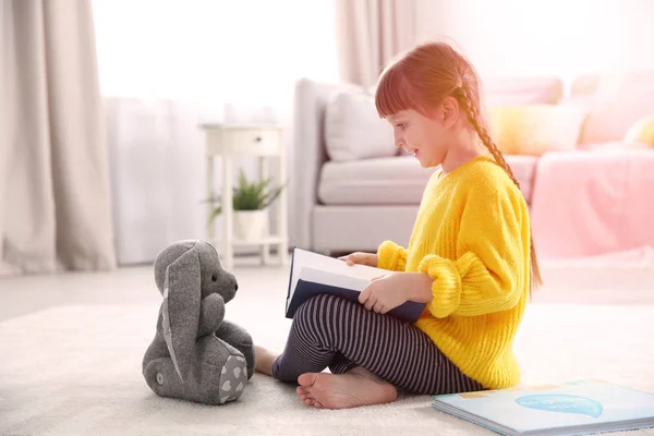 Menina bonito com brinquedo sentado no chão e ler livro em casa — Fotografia de Stock